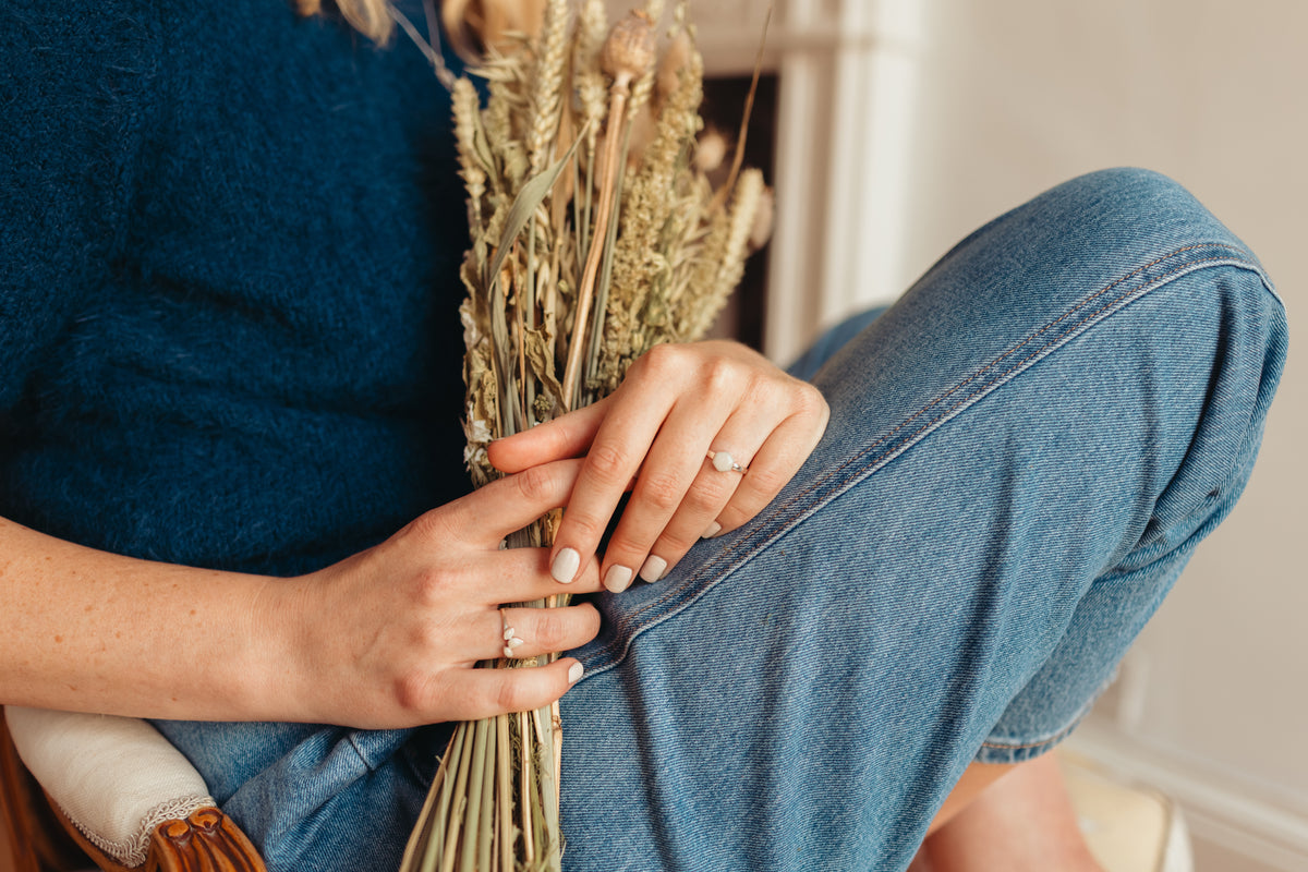A close-up lifestyle shot of a person wearing a dark blue knit sweater and blue jeans, holding a small bunch of dried wheat stems. The person's hands are resting on their knee, showing off silver rings.